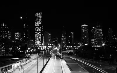 Long exposure headlights streak up a wide freeway toward a monochrome downtown, the near bank of the road left in solid black.