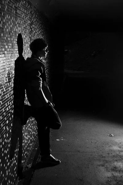 Black and white photo of a young man resting against a lit brick wall, the tunnel ahead of him falling into complete darkness.