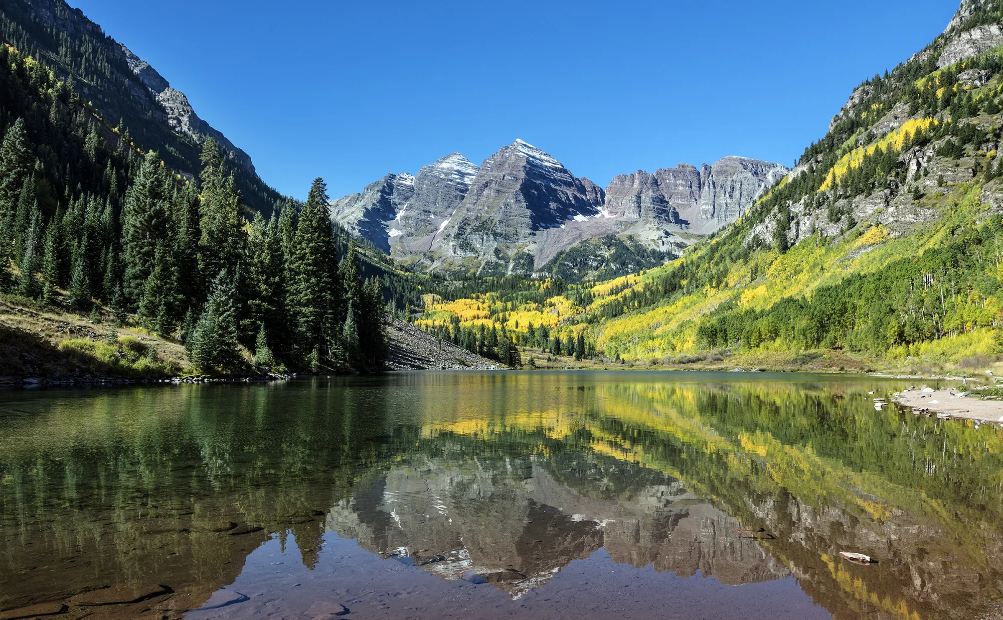 Twin grey peaks mirror in a still lake ringed by pines and yellow autumn aspens, the water doubling the scene across the bottom half.