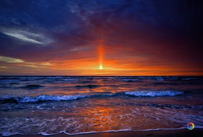 A vertical column of red light rises from the setting sun into slate-blue clouds, choppy waves catching its glow all the way to the sand.