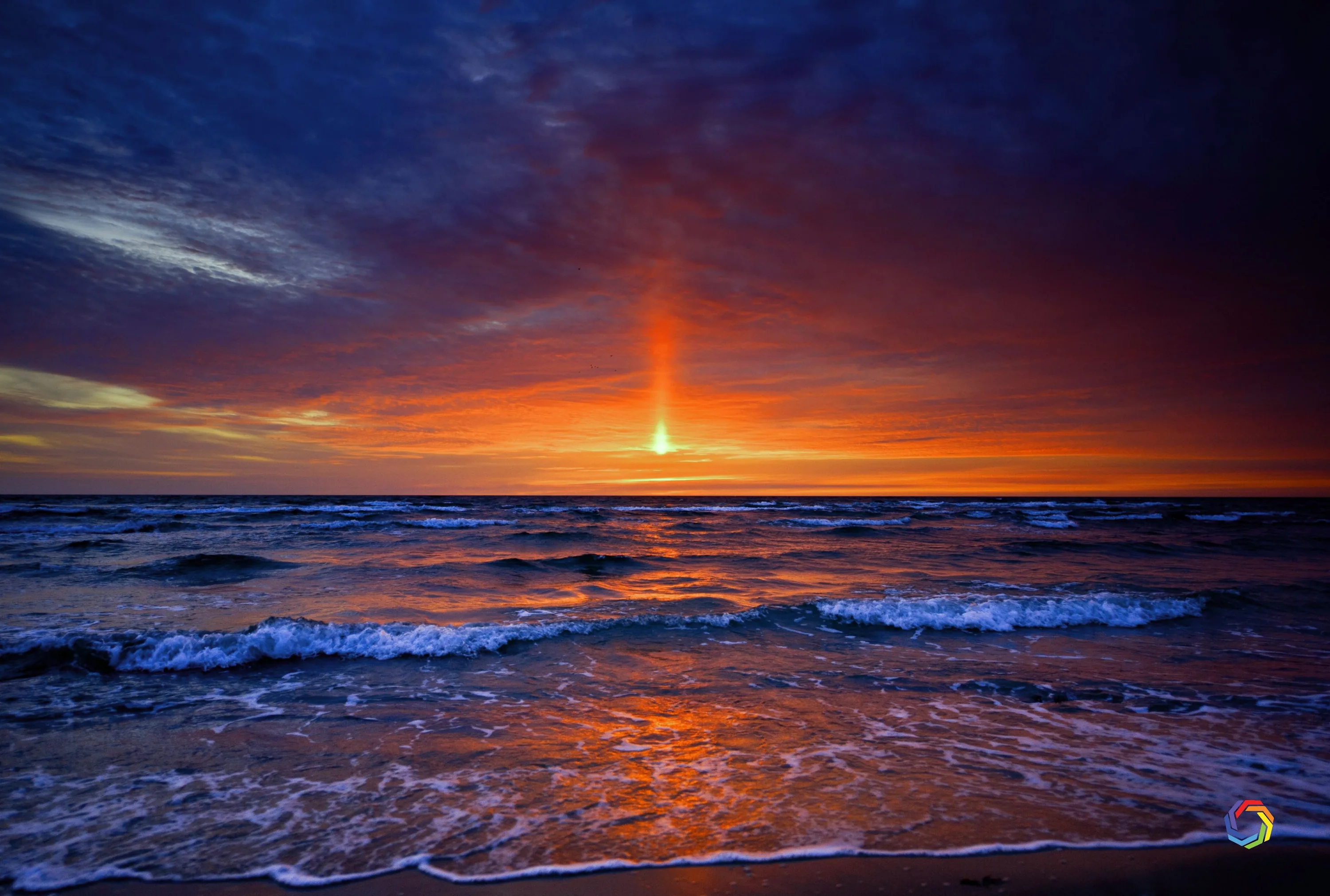 A vertical column of red light rises from the setting sun into slate-blue clouds, choppy waves catching its glow all the way to the sand.