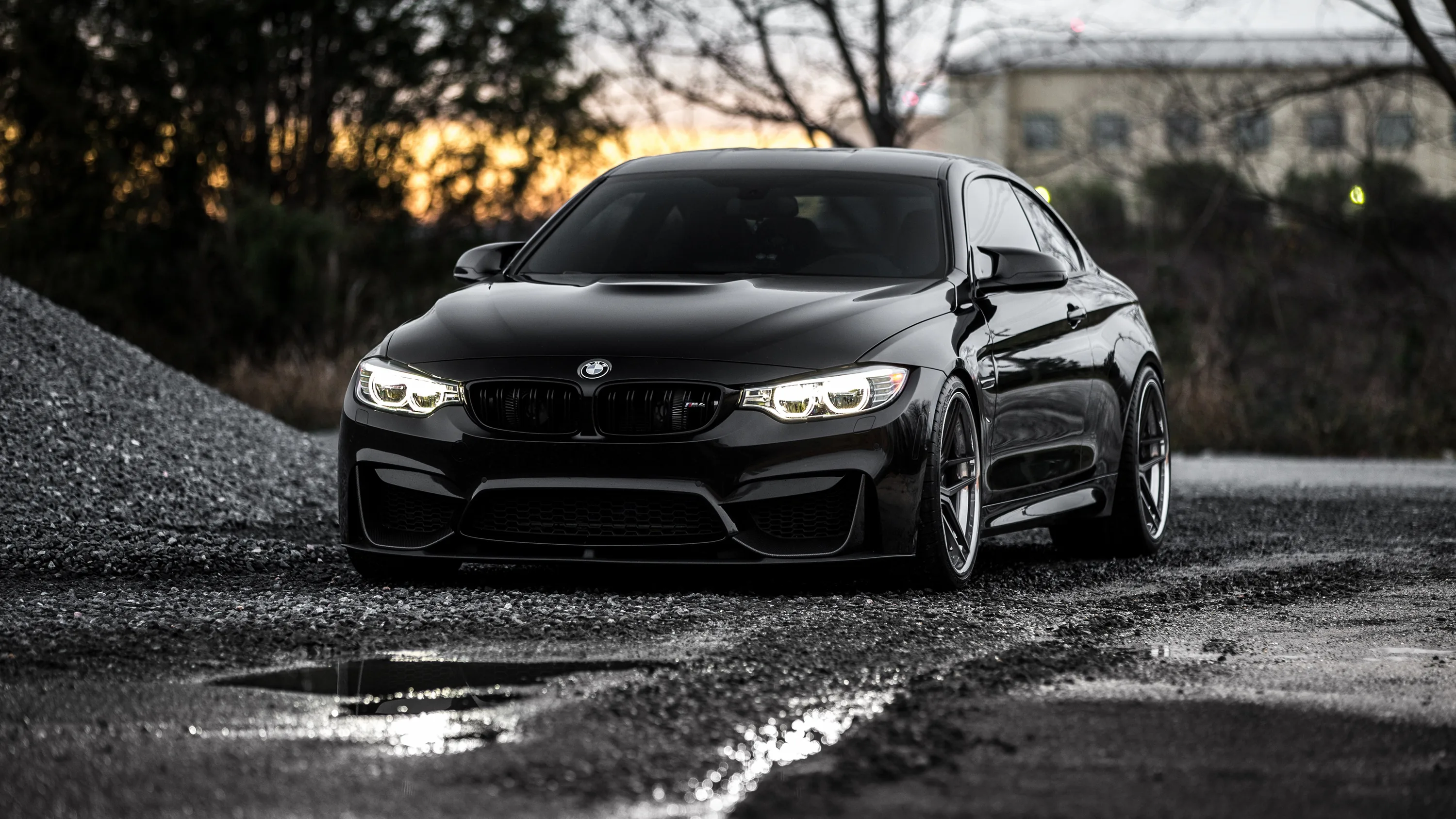 A black BMW coupe sits on wet tarmac at dusk, its ring headlights burning while the sunset behind drops to a thin orange line.