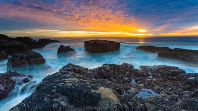 An orange band of sunrise burns behind wave-worn rocks, the long exposure turning the water to fog so only the stones stay sharp.