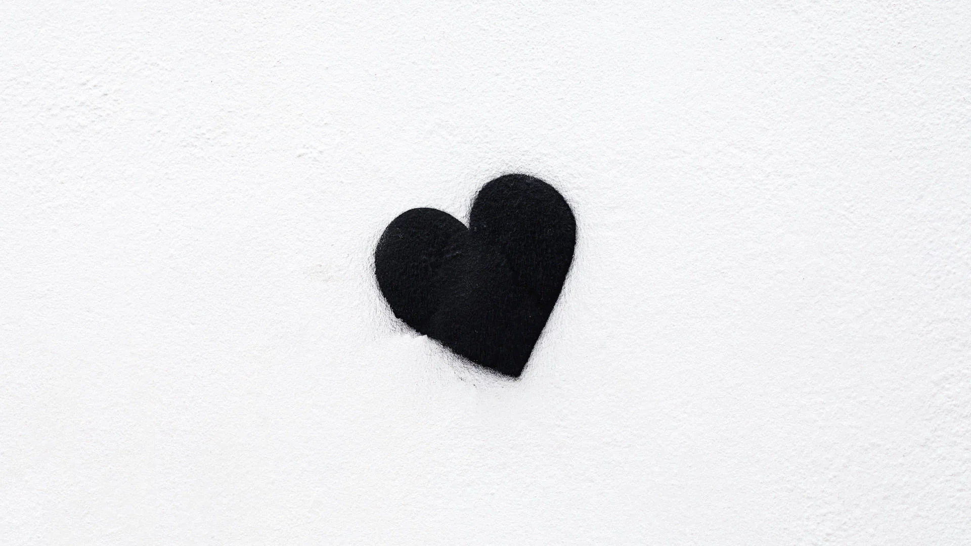 A small hand-painted black heart sits just left of centre on rough white plaster, the rest of the wall left plain and evenly lit.