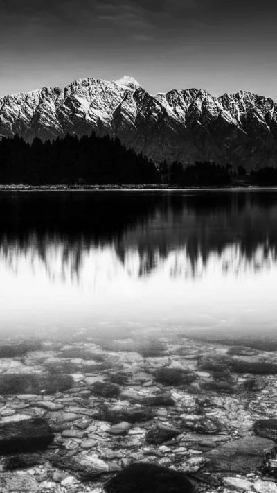 A jagged snow-capped ridge mirrors in flat water, and the shallows in the lower half go clear enough to show every stone on the bottom.