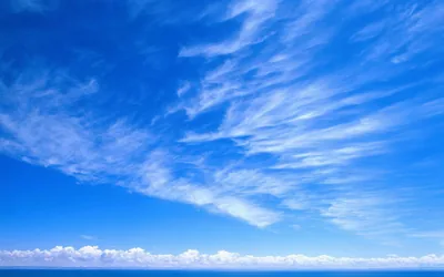 Cirrus streaks fan diagonally across a deep blue sky above a thin strip of sea, the upper right corner left almost clear.