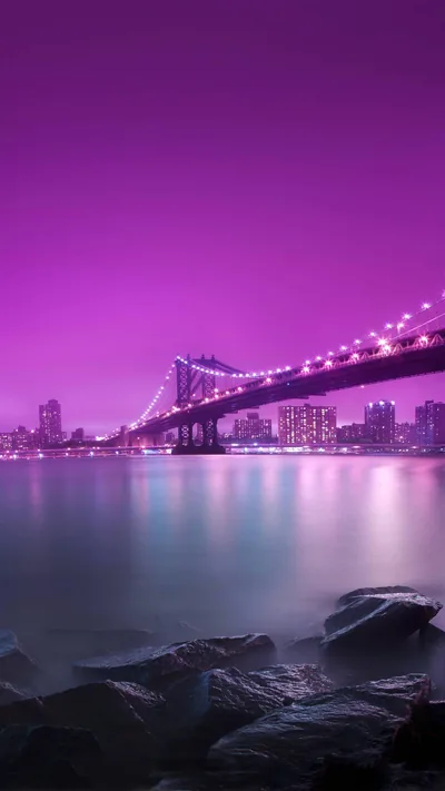 A suspension bridge crosses still water beneath a deep magenta sky, its lights reflecting in long streaks and dark rocks filling the near shore.