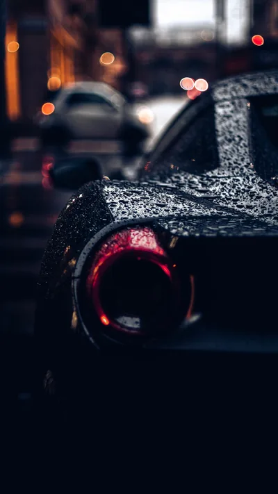 Close on the rear haunch of a wet supercar at night, water sitting in beads across the paint and city lights breaking into orange bokeh behind the round tail lamp.