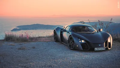 A low-slung matte coupe parked on a dirt clifftop, the sea and a distant headland going pink behind it as the sun drops out of frame.