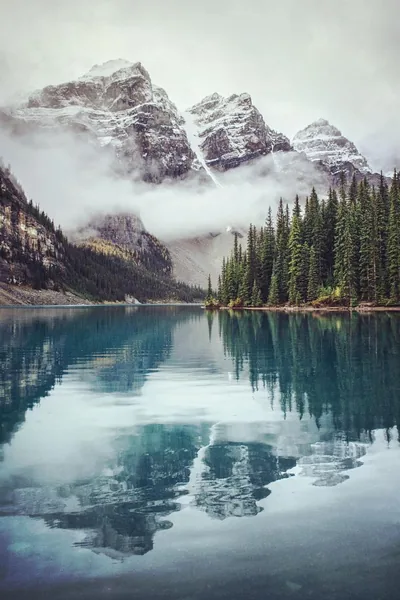 Mist wraps a wall of grey summits behind spruce forest, their reflection sinking into steel-blue water that stays dark enough for lock-screen text.