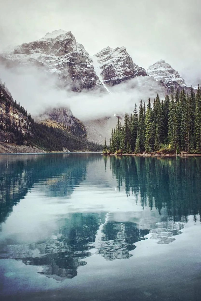 Mist wraps a wall of grey summits behind spruce forest, their reflection sinking into steel-blue water that stays dark enough for lock-screen text.