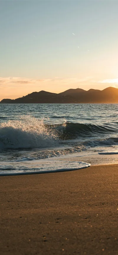 A small wave throws up spray against distant mountains, warm low light grazing the sand that fills the lower third of the frame.