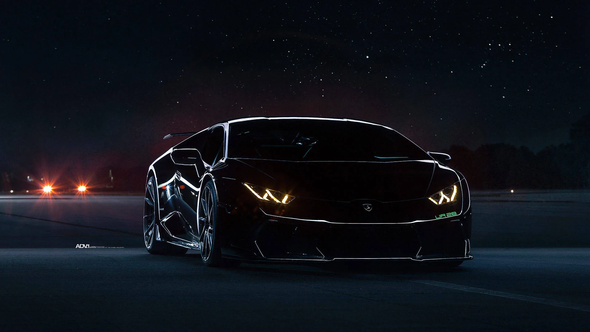 A wide-body Lamborghini faces the camera on an empty road at night, its angular headlights glowing amber while stars and distant orange lights fill the background.