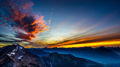 A ragged cloud catches fire in orange over snow-dusted ridges, a thin gold band along the horizon while the valleys below fall into blue shadow.