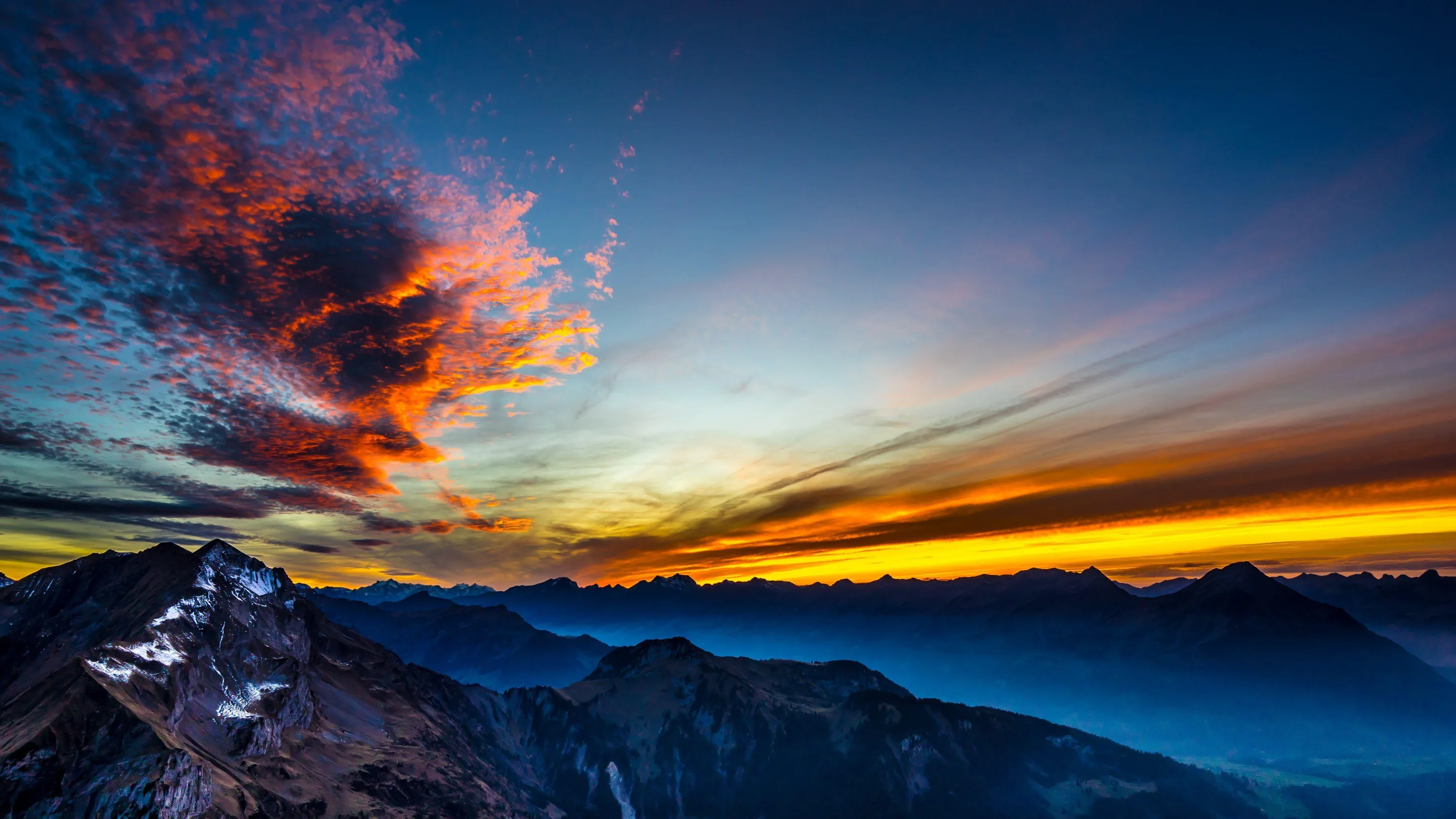 A ragged cloud catches fire in orange over snow-dusted ridges, a thin gold band along the horizon while the valleys below fall into blue shadow.