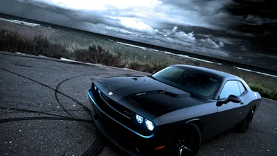 A blacked-out Challenger stands on a cracked shoreline lot ringed by old burnout marks, heavy storm clouds pressing down over the sea behind.