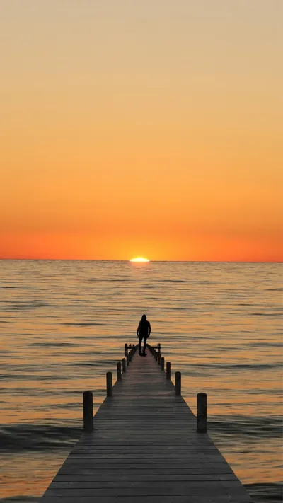 A figure sits at the end of a long wooden jetty facing the last sliver of sun, the plain orange sky above left empty for a lock-screen clock.