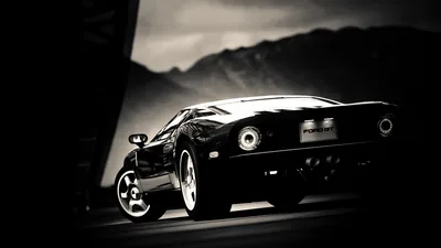 The rear three-quarter of a black sports car sits low against hills and sky, its round tail lights and rims the brightest points in a near-monochrome shot.