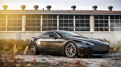 An Aston Martin on bronze forged wheels parked on broken concrete, sun flaring past the corner of a boarded-up factory with red brake calipers showing through the spokes.