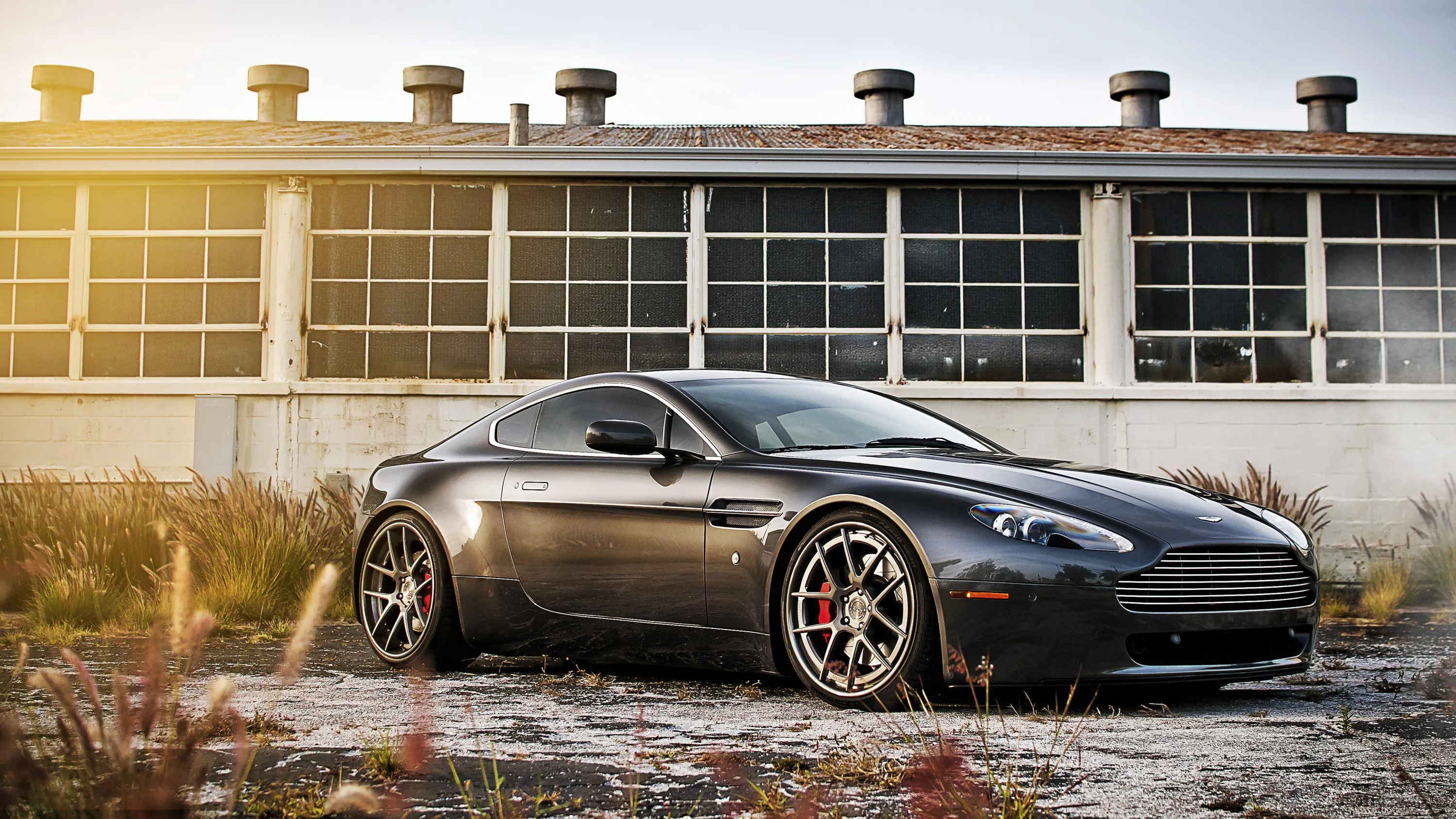An Aston Martin on bronze forged wheels parked on broken concrete, sun flaring past the corner of a boarded-up factory with red brake calipers showing through the spokes.