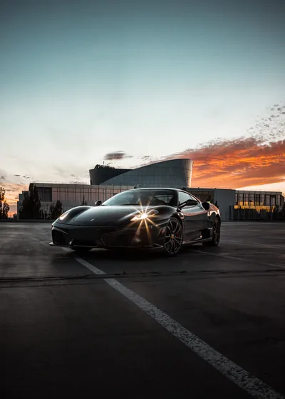 A low black supercar on empty parking asphalt, the last of the sun splitting into a star across its bonnet as orange cloud streaks the horizon.