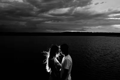 A couple stand forehead to forehead at the edge of a lake, her hair caught by the wind, a heavy bank of cloud filling the sky behind them in monochrome.