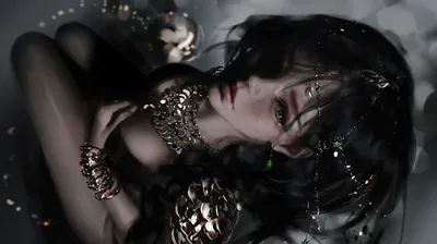 Dark hair spreads across the frame as she rests one hand near her face, gold rings and beads catching the only light in the shot.