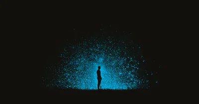 Thousands of glowing blue specks spray outward behind a standing silhouette, and beyond the burst every corner is true black, kind to AMOLED panels.