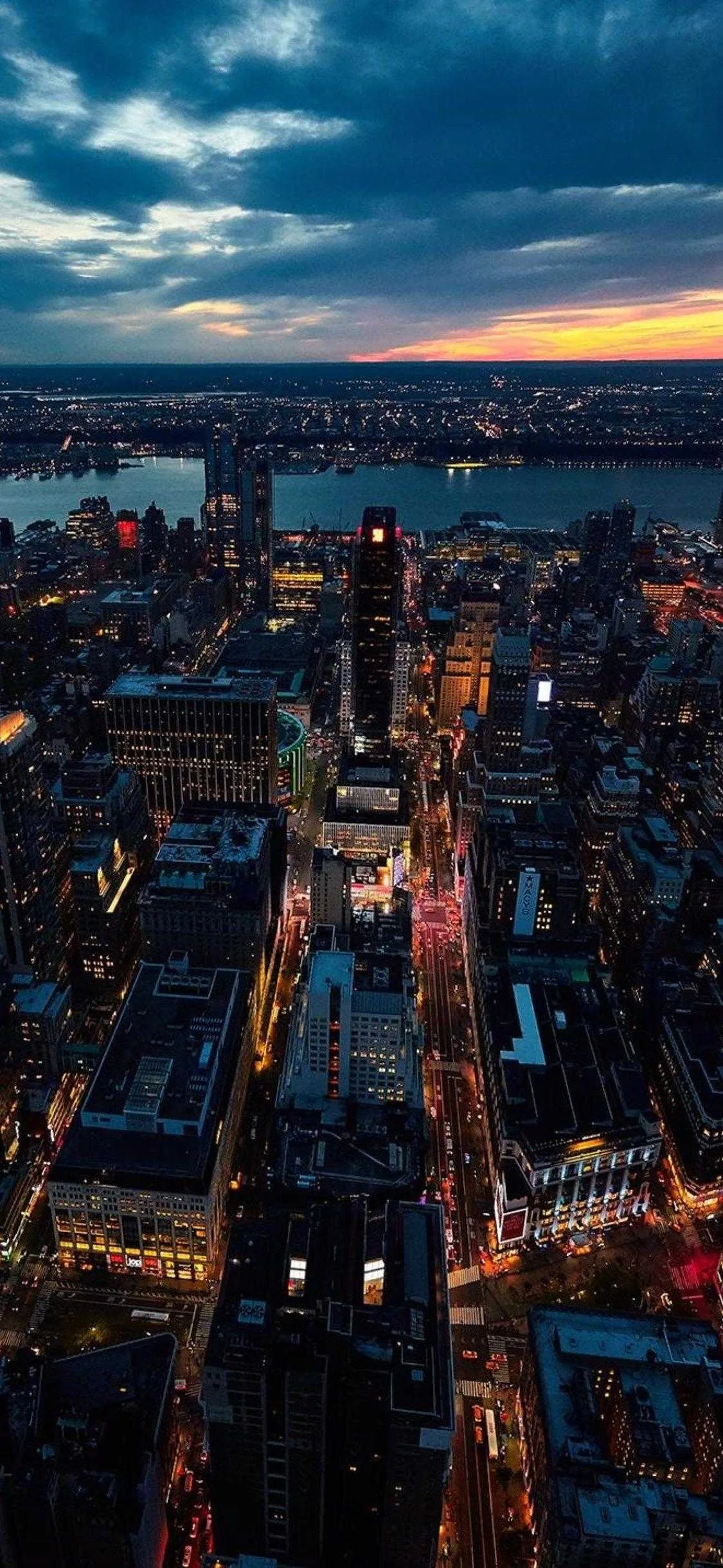 Seen from a skyscraper at dusk, Midtown streets glow in strips of red and gold below sunset clouds, the Hudson fading grey along the top.
