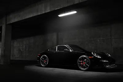 A low 911 waits under a single strip light in a bare concrete garage, red brake calipers showing through the spokes while the left half stays dark.