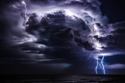 Lightning glows inside a vast storm cell over a night sea, one bolt escaping to the water — the frame nearly monochrome violet throughout.