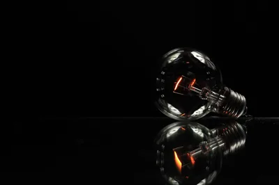 A clear glass bulb lies on its side on a reflective black surface, filament glowing orange, with the whole left half of the frame kept empty.