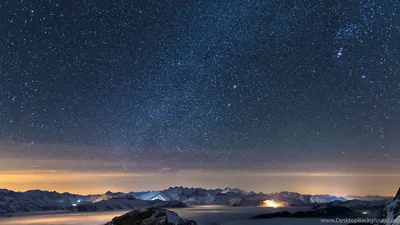 Stars thicken toward the upper left over moonlit ranges, ribbons of village light tracing the valleys along the bottom edge.