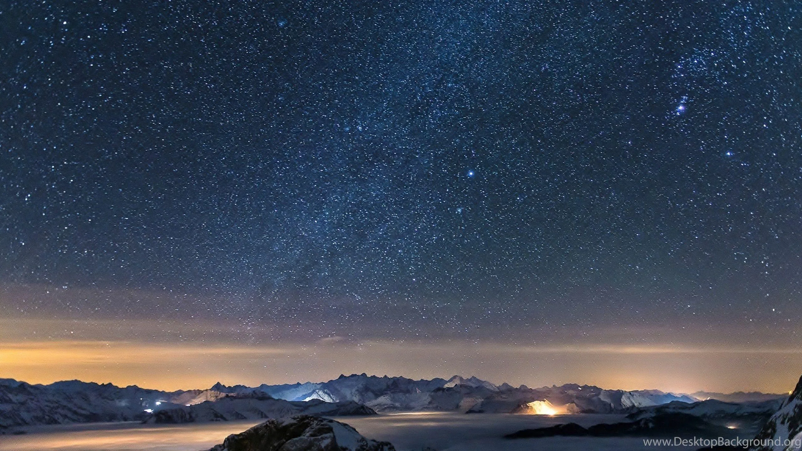 Stars thicken toward the upper left over moonlit ranges, ribbons of village light tracing the valleys along the bottom edge.