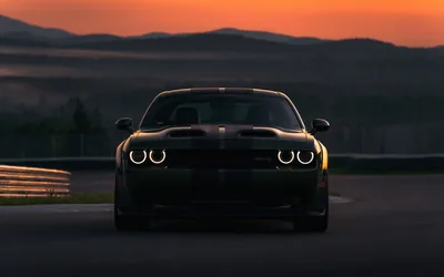 The muscle coupe rolls straight at the camera on an empty circuit, ring-shaped headlamps burning white against an orange band of sunset over dark hills.