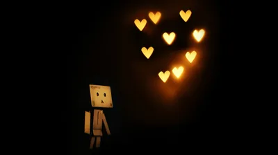 A small cardboard robot stands in the dark looking up at seven warm bokeh hearts floating above its head.