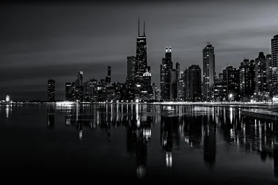 A skyline of tall towers glows along a waterfront at night, its lights repeated in the calm surface below, all in cool monochrome.