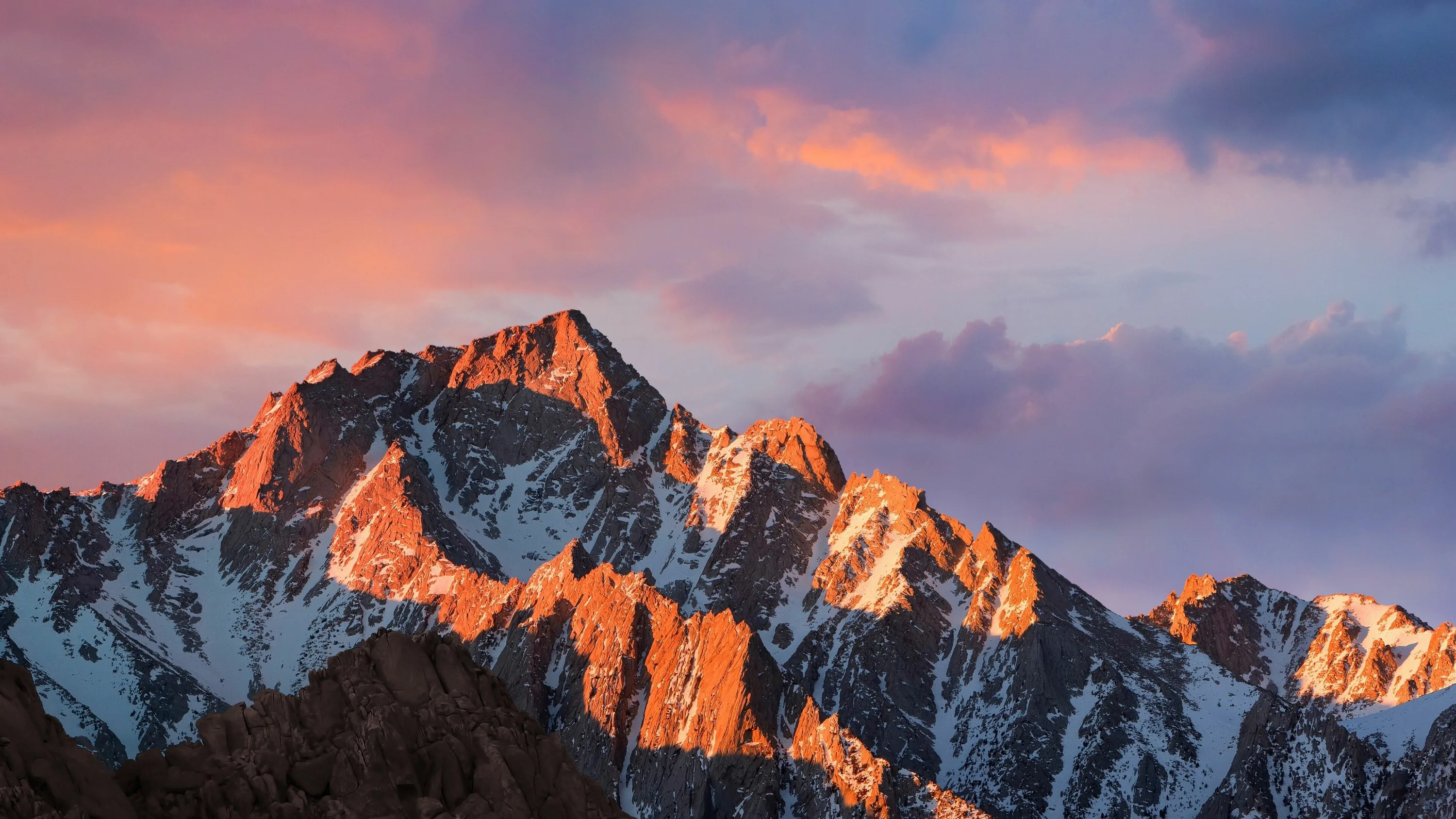 Last light turns a jagged granite ridge orange-pink under drifting violet clouds, in the style of the macOS Sierra desktop.
