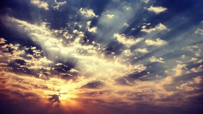 Crepuscular rays burst from a low sun through dark broken cloud, the light pooling in the lower left while the corners fall to near black.