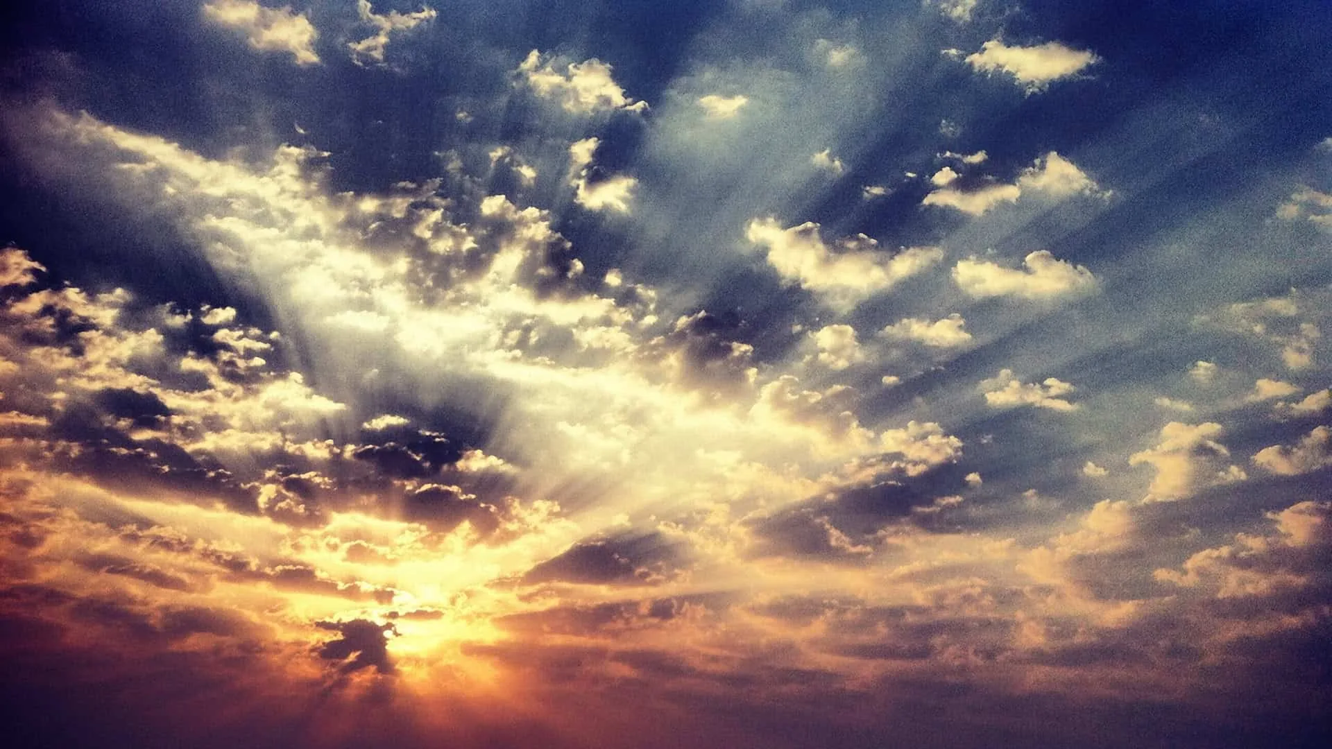 Crepuscular rays burst from a low sun through dark broken cloud, the light pooling in the lower left while the corners fall to near black.