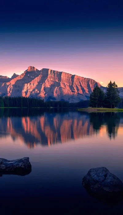 A pink-lit rock face mirrors perfectly in flat water, with two boulders breaking the surface at the bottom and the sky deepening to navy overhead.