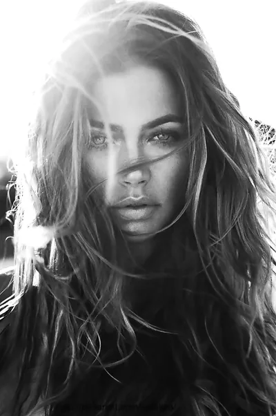Sunlight flares from the upper left behind a woman's loose windblown hair in black and white, her shaded face at centre and the lower third falling into shadow.