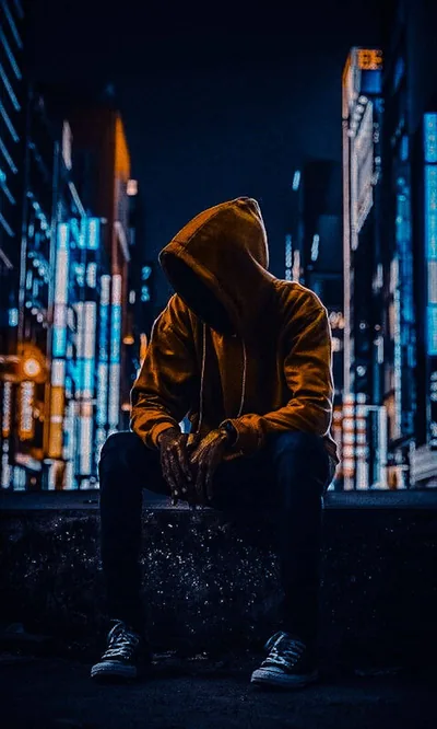 Head bowed in a mustard hoodie and sneakers, he sits on a concrete edge with the blue bokeh of skyscrapers stretched behind him.
