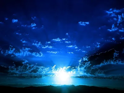 White light breaks over a low horizon and throws rays up through heavy blue cloud, the land at the bottom reduced to a dark strip.