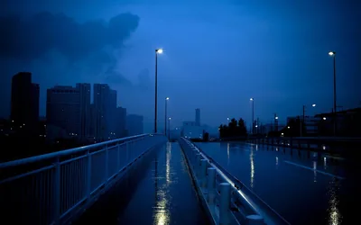 A wet footbridge runs toward a city skyline at blue hour, lamp posts reflecting on the pavement, the sky's upper left heavy with dark cloud.