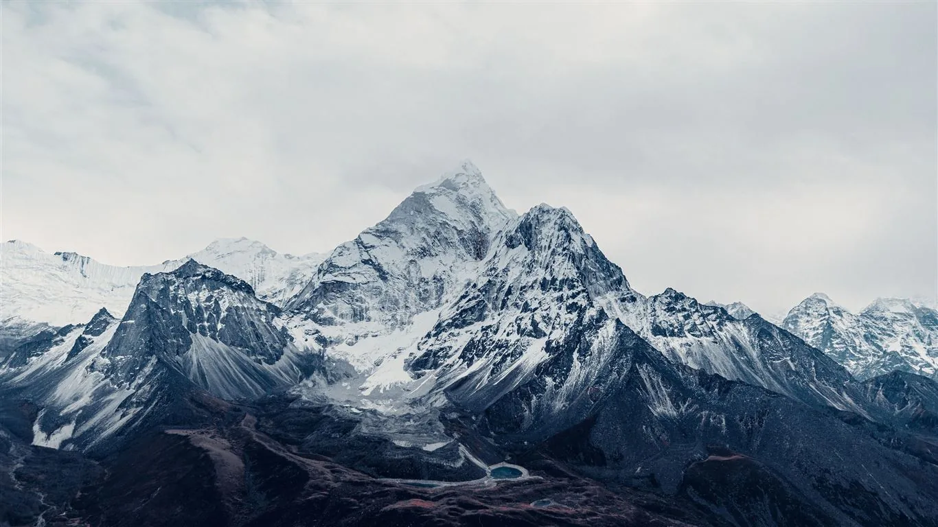 A steep-sided Himalayan summit stands among wind-scoured ridges, its faces streaked black and white beneath a flat grey sky in near-monochrome.