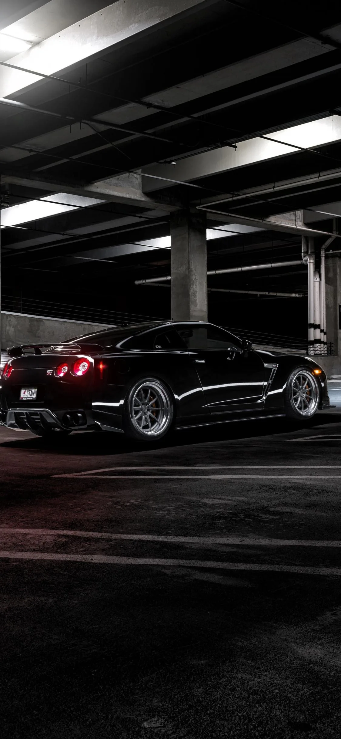 Vertical shot of a black Mustang on the empty top deck of a car park, red tail lamps lit and strip lighting running away along the ceiling.