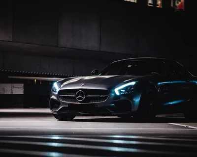 A grey Mercedes-AMG GT rolls through a dark parking structure at night, its headlights casting blue light across the striped road paint below.