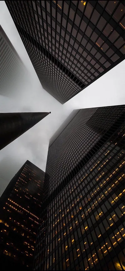 Dark office towers lean into an overcast sky shot straight up, lit windows dotting the facades while the pale centre leaves room for a clock.