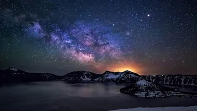 The Milky Way tilts in purple and violet over a snow-rimmed crater lake, an island silhouette at the waterline and orange glow leaking along the horizon.